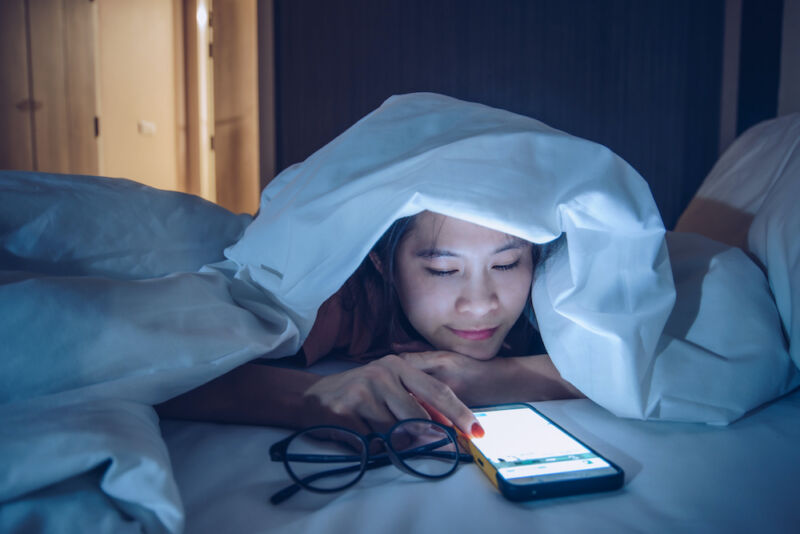 A woman under a thick blanket looks at her smartphone.